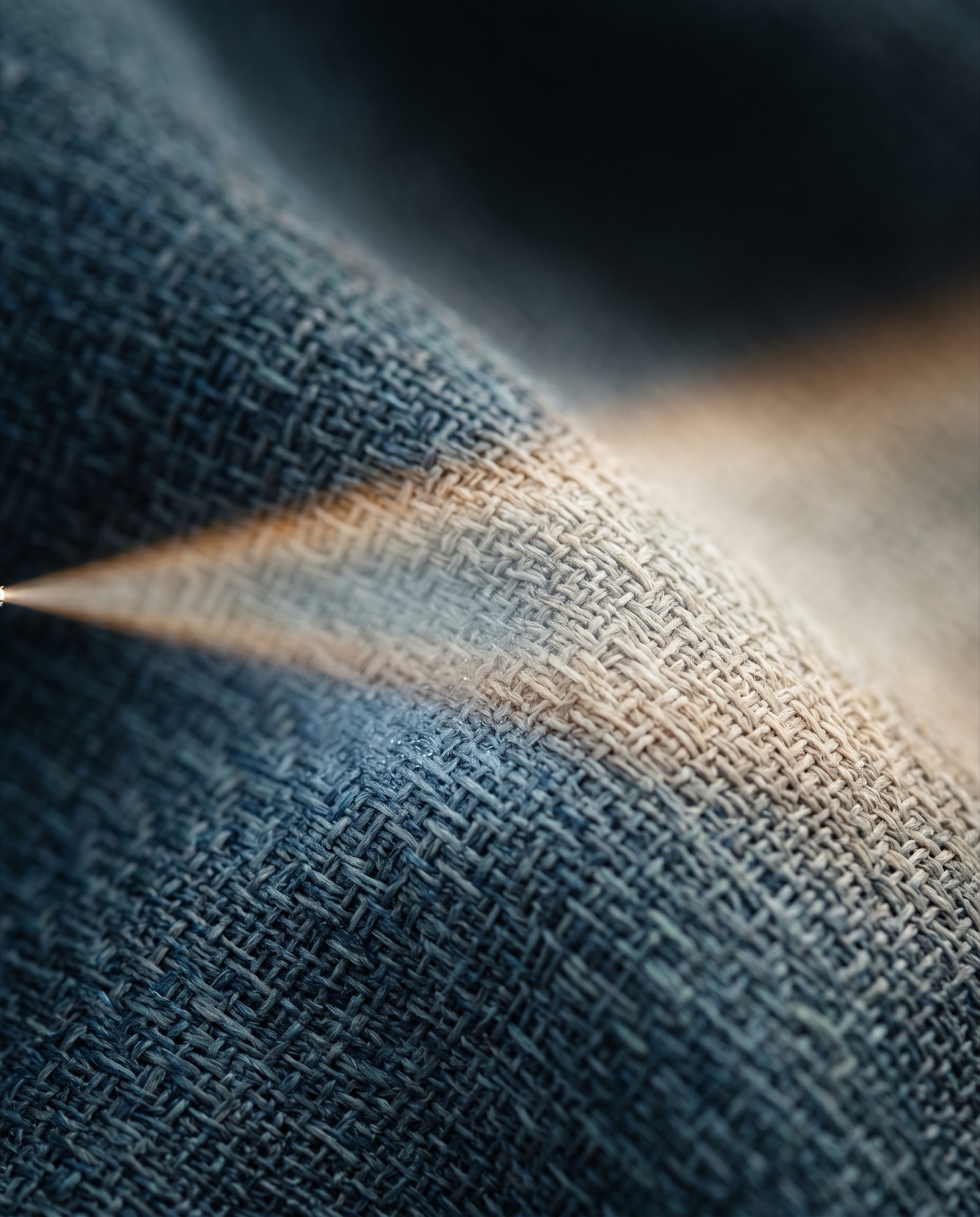 Macro of dusty blue linen draped in a soft diagonal fold, a narrow beam of warm golden light on the weave against cool shadow