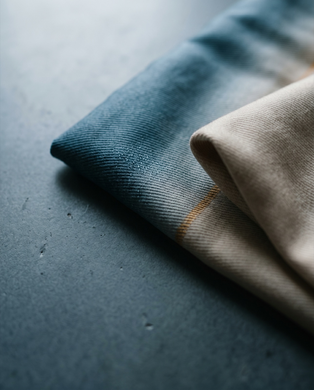Folded bedding fabric with a gradient from deep teal-blue through a thin golden stripe to warm beige on dark stone