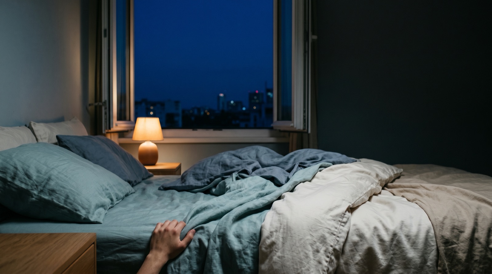 A dimly lit bedroom at night with a warm bedside lamp and an open window revealing distant city lights